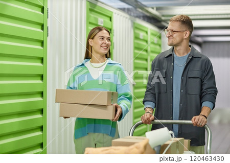 Medium shot of cheerful woman carrying cardboard boxes walking along self storage warehouse hallway with helping boyfriend pushing dolly cart during apartment decluttering 120463430