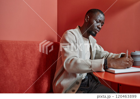 Side view of hard working African American businesswoman writing in notebook planning schedule ahead while sitting at cafe table with colorful minimalistic design, copy space 120463899
