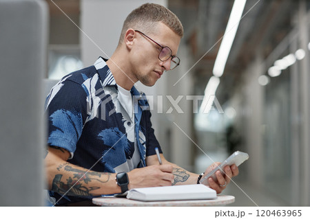 Side view of young male business worker checking message on mobile phone writing in notebook while sitting at table in common area of modern office building, copy space 120463965
