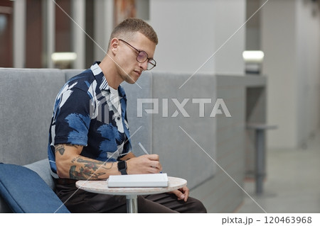 Side view of young male business worker concentrated on writing in notebook sitting at table in common area of modern office building, copy space 120463968