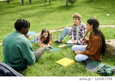 Group of students sitting on grassy field, discussing study topics together. Engaging academic dialogue, surrounded by nature and open books on vibrant green grass 120467704