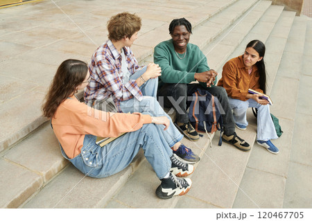 Four friends of diverse backgrounds smiling while sitting on steps. Group chatting and enjoying time together with smiles and camaraderie among the mixed-ethnic group 120467705