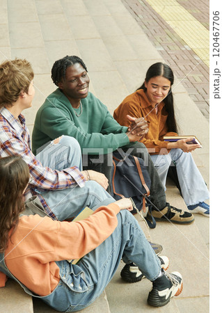 Group of four diverse friends sitting on steps in outdoor urban area, engaged in friendly conversation and sharing stories, holding books and wearing casual clothes 120467706