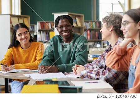Group of students sitting at table in library, engaging in study session and discussion. Background includes bookshelves and study materials, adding to academic atmosphere 120467736