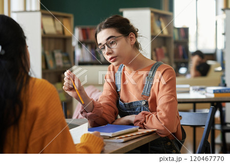 Caucasian young woman wearing glasses and overalls talking with friend while studying in library setting with bookshelves in background 120467770