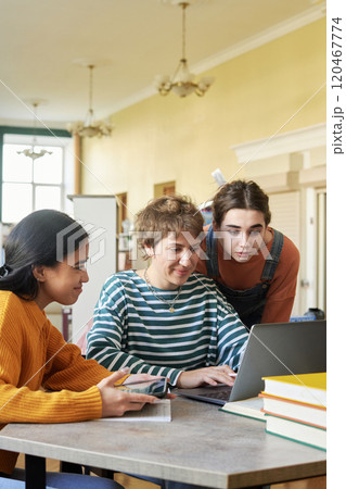 Three young students working together on homework in a bright room. Engaging with each other while using laptop and taking notes with books stacked on table Three young students working together on homework in a bright room. Engaging with each other while using laptop and taking notes with books stacked on table 120467774