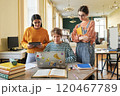 Three students engaging in collaborative study session in a spacious room with books, laptops, and tablets. Diverse group concentrating on assignments while interacting with each other 120467789