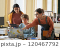 Three students are collaborating on a group project while using a laptop and tablet in classroom environment with bookshelves and windows 120467792