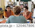 Group of students sitting at table discussing study materials and sharing ideas in library during study session, with books and shelves in background 120467806