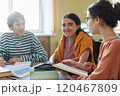 Group of friends collaborating on study session in library, sitting around table with books and notebooks, engaged in discussion and smiling 120467809