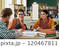 Three students sitting in library discussing class project while surrounded by books and educational materials where one person smiling warmly and the others listening attentively 120467811