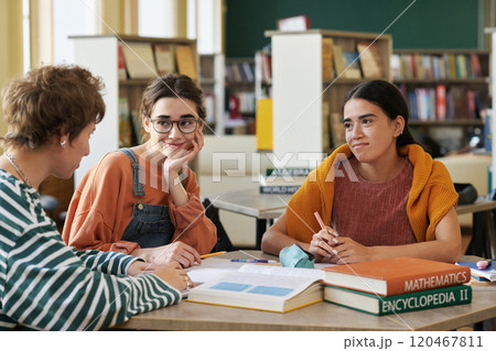 Three students sitting in library discussing class project while surrounded by books and educational materials where one person smiling warmly and the others listening attentively Three students sitting in library discussing class project while surrounded by books and educational materials where one person smiling warmly and the others listening attentively 120467811