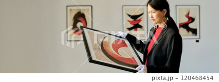 Woman in modern art gallery examining vibrant abstract art while standing with framed artwork in hands, showcasing engagement with contemporary art 120468454