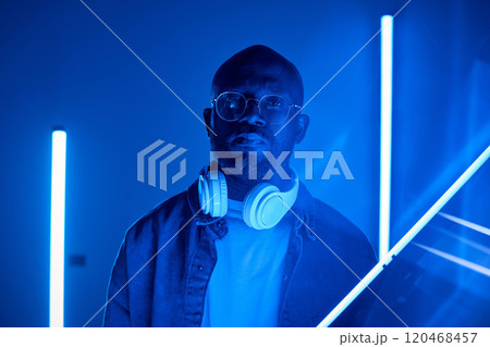 Portrait of a man looking straight at the camera, wearing headphones under neon blue light, creating an atmospheric scene. The setting includes vertical light bars adding to the futuristic ambience Portrait of a man looking straight at the camera, wearing headphones under neon blue light, creating an atmospheric scene. The setting includes vertical light bars adding to the futuristic ambience 120468457