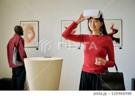 Portrait of young woman engaging with virtual reality headset in modern art gallery with abstract paintings and another visitor in the background Portrait of young woman engaging with virtual reality headset in modern art gallery with abstract paintings and another visitor in the background 120468496