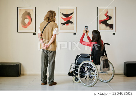 Two people in modern gallery observing and photographing abstract art pieces displayed on wall. One person is standing while other is sitting in a wheelchair Two people in modern gallery observing and photographing abstract art pieces displayed on wall. One person is standing while other is sitting in a wheelchair 120468502