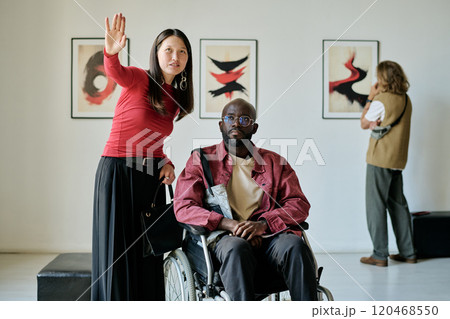 Woman in red shirt gesturing while standing next to man in wheelchair in art gallery with background of framed abstract paintings and another person observing Woman in red shirt gesturing while standing next to man in wheelchair in art gallery with background of framed abstract paintings and another person observing 120468550