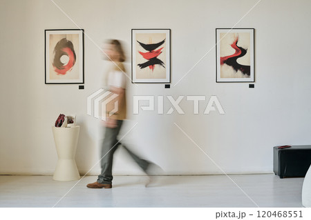 Blurred figure walking through art gallery exhibiting abstract paintings on wall demonstrating motion. Artwork showcasing dynamic brushstrokes and vivid contrasts 120468551