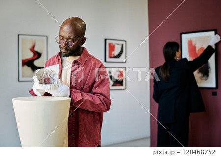 Man holding sculpture and woman hanging painting in contemporary art gallery while engaging with displays and creations from artists on exhibition 120468752