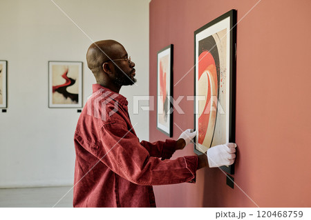 African American man examining abstract artwork hanging on wall in art gallery setting displaying white gloves and red shirt African American man examining abstract artwork hanging on wall in art gallery setting displaying white gloves and red shirt 120468759