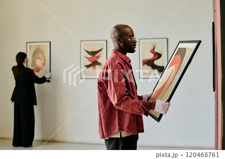 African American man holding abstract painting, curating art gallery exhibit. Background reveals another person examining framed artwork on white walls 120468761