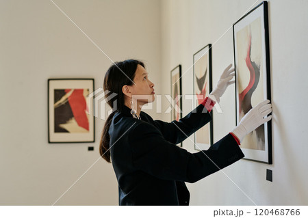 Woman adjusting framed artwork on gallery wall wearing gloves. Modern abstract paintings hanging alongside for exhibition Woman adjusting framed artwork on gallery wall wearing gloves. Modern abstract paintings hanging alongside for exhibition 120468766