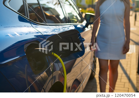 Woman in a white dress walks towards an electric car at a charging station during daylight 120470054