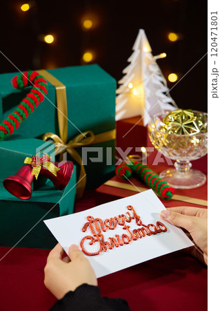 Female's hands holding a christmas card on red covered table, next to it is variety christmas decorative things like gift boxes, christmas balls, bell, pine and sparkling yellow led lights in back Female's hands holding a christmas card on red covered table, next to it is variety christmas decorative things like gift boxes, christmas balls, bell, pine and sparkling yellow led lights in back 120471801