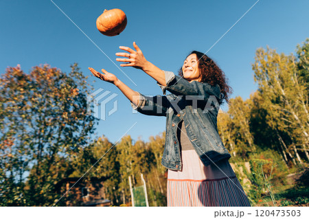 Woman with pumpkin on a sunny day, embracing the harvest season, Rural life in fall. Woman playing with a pumpkin in her garden, celebrating the bounty of autumn Woman with pumpkin on a sunny day, embracing the harvest season, Rural life in fall. Woman playing with a pumpkin in her garden, celebrating the bounty of autumn 120473503