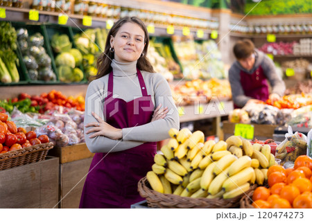 Young female seller standing by basket full of bananas in grocery market Young female seller standing by basket full of bananas in grocery market 120474273