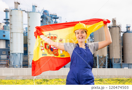 National flag of Spain in the hands of girl in overalls against background of modern metallurgical plant 120474503