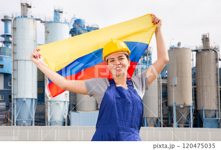 Positive young female worker in helmet waving national flag of Colombia while standing in front of big tanks at refinery factory on sunny summer day 120475035