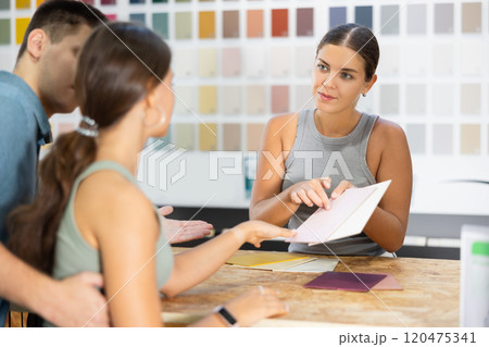 Female salesperson in paint and varnish department is assisting couple customers by showcasing product samples and providing guidance through product catalog at counter 120475341