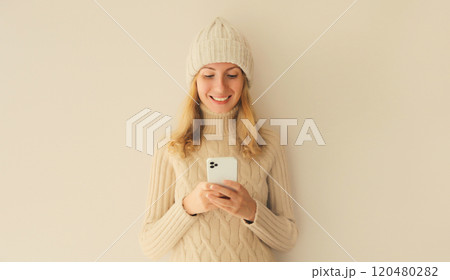 Portrait happy smiling young woman with phone in winter hat, white sweater, girl holding smartphone 120480282