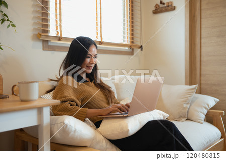 Smiling happy young woman wearing a warm brown sweater sitting on sofa with laptop Smiling happy young woman wearing a warm brown sweater sitting on sofa with laptop 120480815