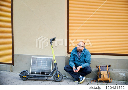 Male person waits while charging electric scooter with photovoltaic solar panel in urban settings. Integration of solar power as sustainable energy source, promoting eco-friendly urban transportation. Male person waits while charging electric scooter with photovoltaic solar panel in urban settings. Integration of solar power as sustainable energy source, promoting eco-friendly urban transportation. 120481522