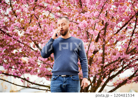 Man allergic using medical nasal drops, suffering from seasonal allergy at spring in blossoming garden. Handsome man treating runny nose in front of blooming tree outdoors. Spring allergy concept. 120481534