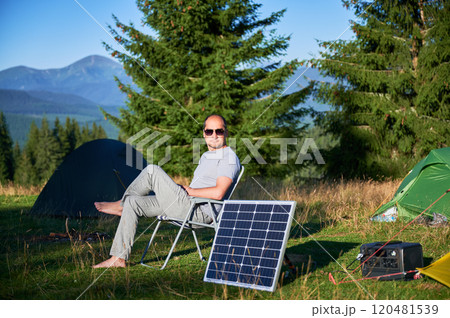 Man using photovoltaic solar panel for charging portable power station outdoor in eco-friendly camping. Male tourist sits relaxed in chair near tents. On background tall pine trees and clear blue sky. Man using photovoltaic solar panel for charging portable power station outdoor in eco-friendly camping. Male tourist sits relaxed in chair near tents. On background tall pine trees and clear blue sky. 120481539