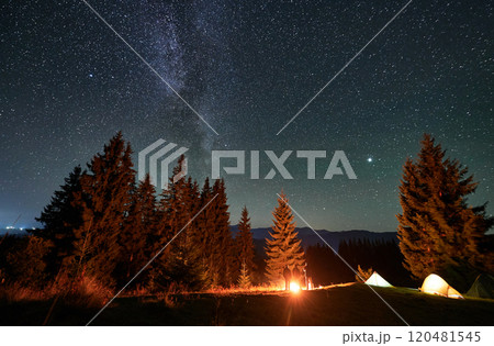 Panoramic view of mountain hill with illuminated camp tents, coniferous trees and hikers under starry sky. Night camping in mountains under beautiful sky with stars. Concept of hiking and traveling. Panoramic view of mountain hill with illuminated camp tents, coniferous trees and hikers under starry sky. Night camping in mountains under beautiful sky with stars. Concept of hiking and traveling. 120481545