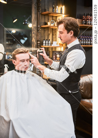 Professional barber using hairspray to finish hairstyle. Stylish hairdresser making final touches. Young customer getting fresh haircut. 120481559
