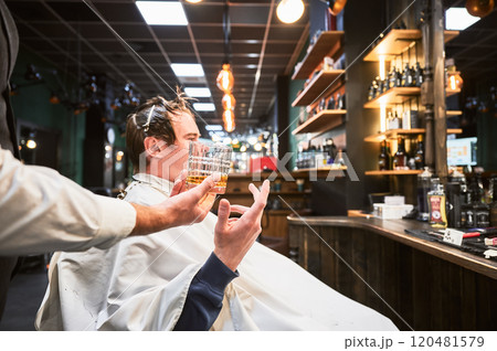 Exclusive offer of drinks from luxury barbershop. Young client taking a glass of whiskey from hairdresser. Side view of male customer getting new haircut. 120481579