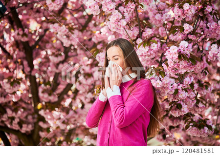 Woman allergic suffering from seasonal allergy at spring in blossoming garden at springtime. Woman sneezing and blowing nose using nasal handkerchief in front of blooming tree. Spring allergy concept. Woman allergic suffering from seasonal allergy at spring in blossoming garden at springtime. Woman sneezing and blowing nose using nasal handkerchief in front of blooming tree. Spring allergy concept. 120481581