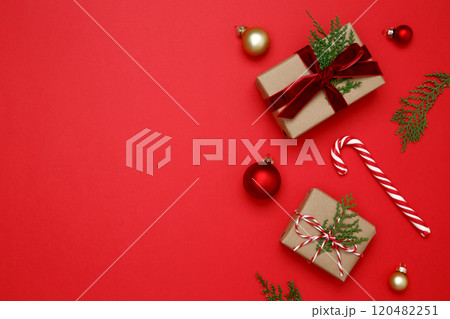 Gifts wrapped in craft paper, Christmas ornaments, thuja branches, and candy cane arranged on red background Gifts wrapped in craft paper, Christmas ornaments, thuja branches, and candy cane arranged on red background 120482251