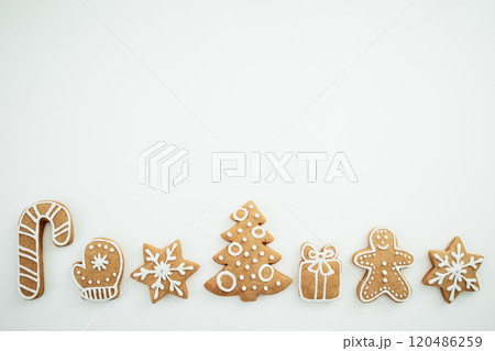 Row of festive gingerbread cookies decorated with white icing, including a candy cane, mitten, snowflakes, Christmas tree, gift box, and gingerbread man, arranged on a white. 120486259