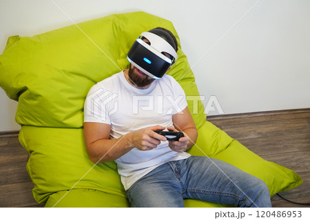 Man leaning back on green bean bag, wearing VR headset, and holding gaming controller Man leaning back on green bean bag, wearing VR headset, and holding gaming controller 120486953