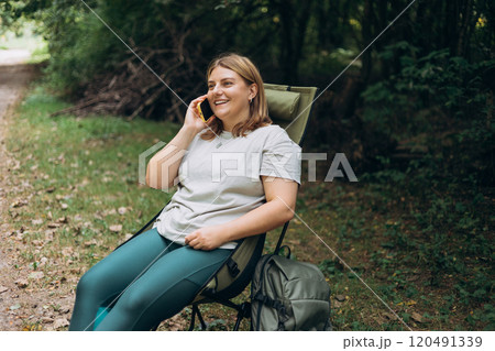 Relaxed happy 30s woman sitting on deck chair in forest and have conversation on a mobile phone, she is planning new travel. Camping, technology and travel concept. A young 30s woman explores a forest Relaxed happy 30s woman sitting on deck chair in forest and have conversation on a mobile phone, she is planning new travel. Camping, technology and travel concept. A young 30s woman explores a forest 120491339