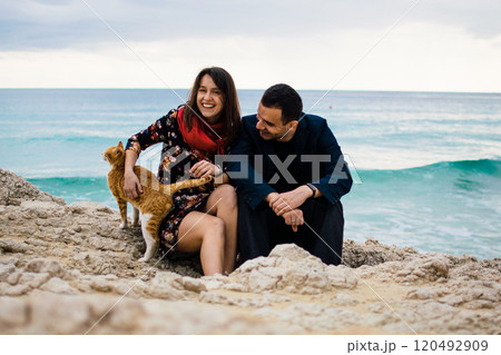 a young couple in love playing with orange stray cat on fabulous rocky coast with turquoise water Ayia Napa, Cyprus 120492909