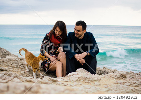 a young couple in love playing with orange stray cat on fabulous rocky coast with turquoise water Ayia Napa, Cyprus 120492912