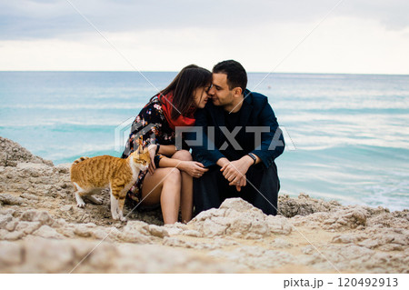 a young couple in love playing with orange stray cat on fabulous rocky coast with turquoise water Ayia Napa, Cyprus 120492913