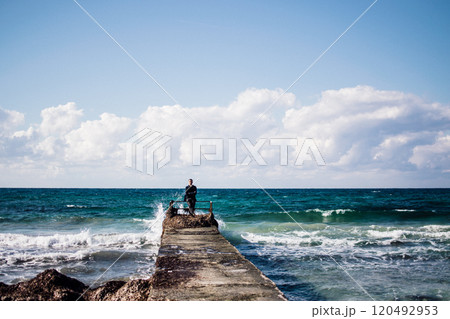 a portrait of a young man on a pier, waves splashing and crashing against the pier 120492953